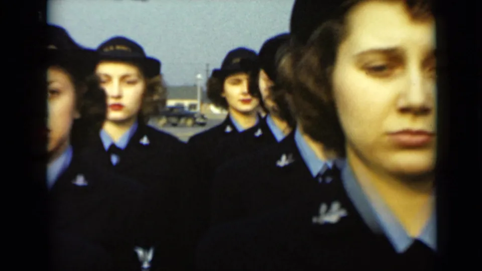 Women in matching dark uniforms and hats stand in rows outdoors, with faces visible and buildings in the distant background.