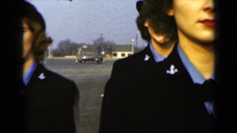 Cropped view of uniformed women in formation outdoors in New York, with low buildings and a vintage car visible in the distance.