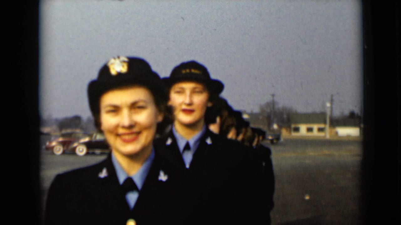 Two uniformed women in dark hats and jackets walk toward the camera on a paved lot.