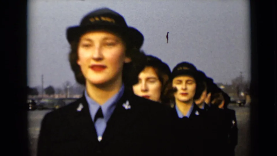 A line of women in dark uniforms and hats moves past the camera in a 1947 New York home-movie shot, with parked cars beyond.