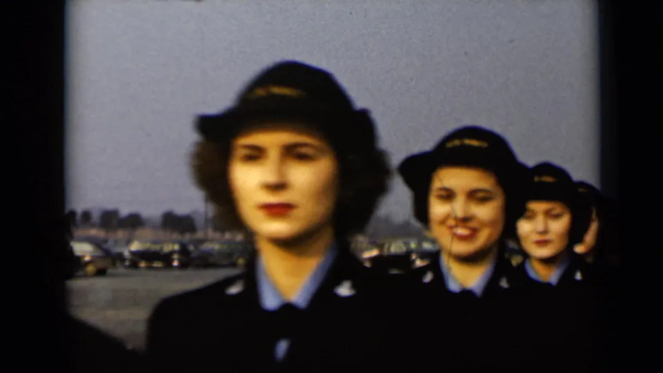 Several women in dark service uniforms and hats walk in single file across a paved lot in 1947 New York, filmed in soft 8mm color.