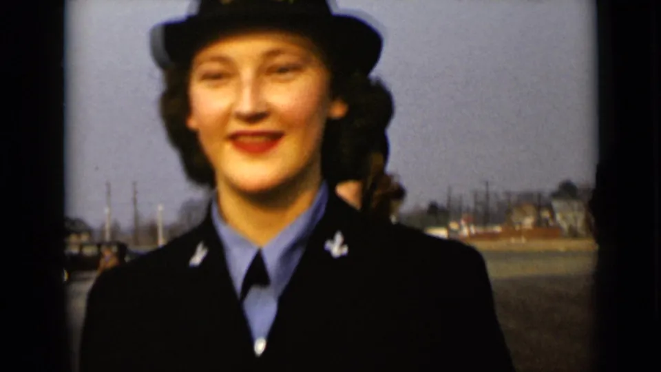 1947 New York home-movie frame of a smiling woman in Navy-style dress uniform, filmed close up in a paved outdoor area.
