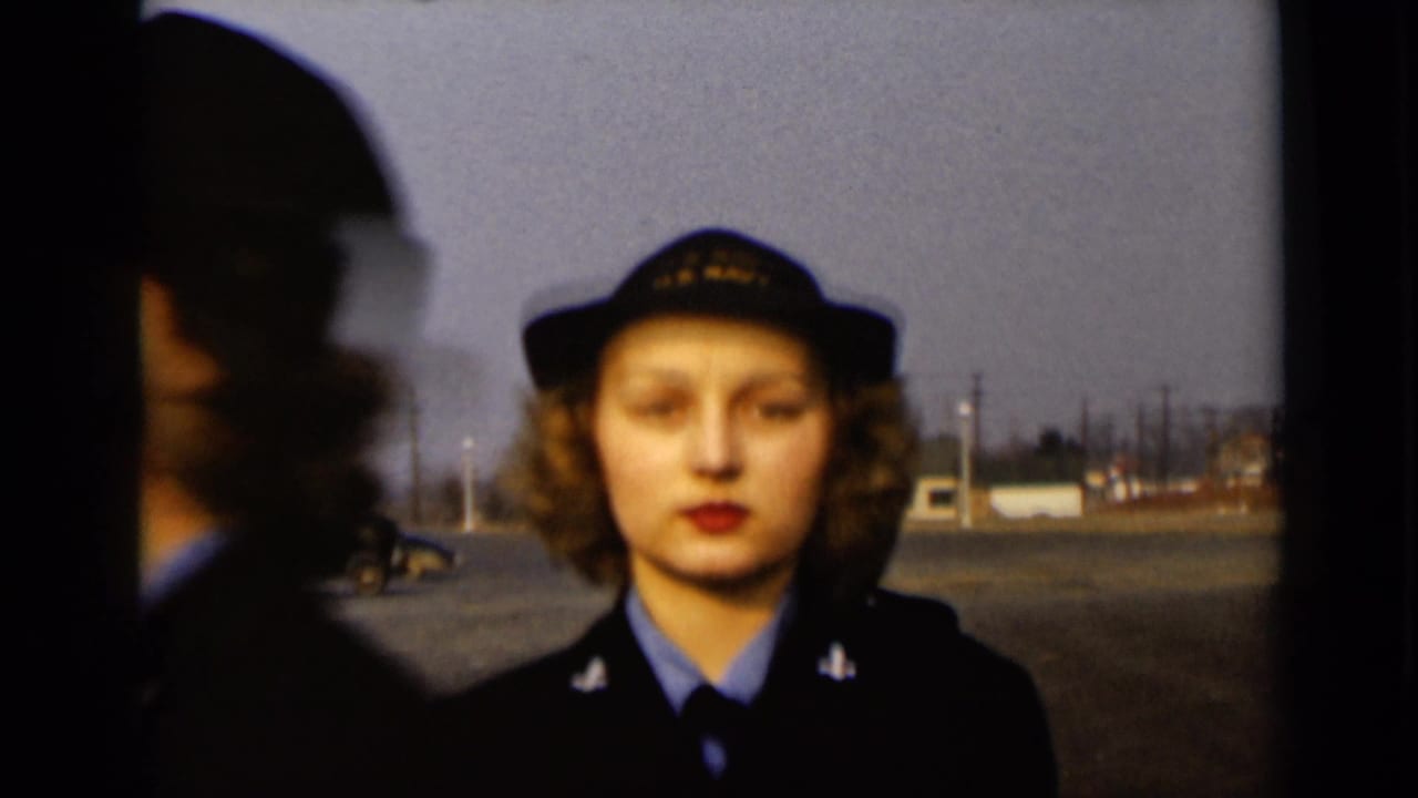 A woman in a U.S. Navy cap and dark jacket faces the camera in New York, with another figure blurred at the edge of frame.