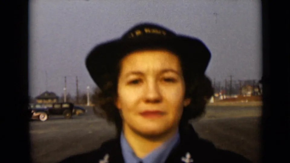 In the 1947 New York home movie, a uniformed woman appears close to camera with a dark period automobile in the background.