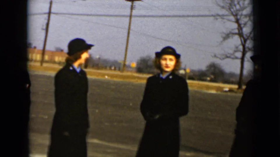 Two women in dark uniforms stand on the paved ground, one turned sideways and one facing the camera, with bare trees and utility poles visible.