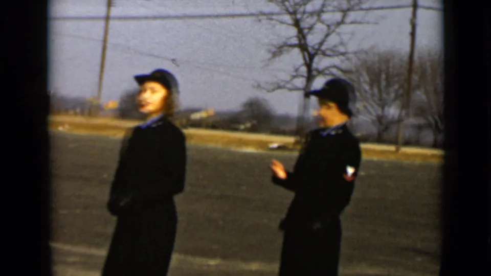 Two women in dark uniforms stand outdoors in profile and partial profile, with a bare tree, open pavement, and distant structures behind them.