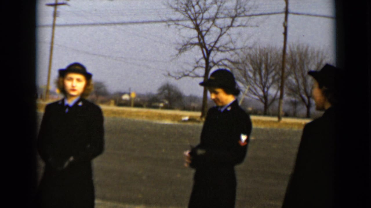 Two women in dark uniforms and hats stand on a quiet street with another figure partly visible.