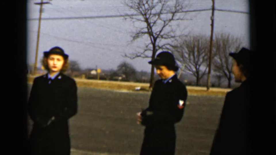 In 1947 New York, women in matching dark uniforms and hats stand outdoors near utility poles and bare trees.