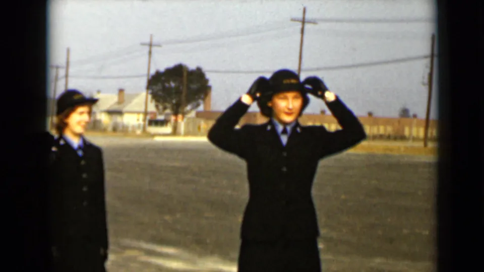 A woman in a dark uniform raises both hands to adjust her hat as another uniformed woman looks on in New York.
