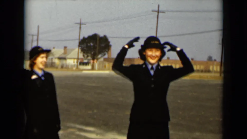 Two women in matching uniforms smile outdoors, with one playfully holding both hands near her hat.
