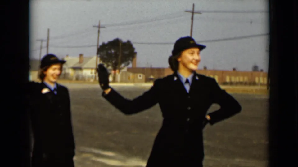 A smiling woman in a dark uniform and hat waves toward the camera on a New York street in 1947.