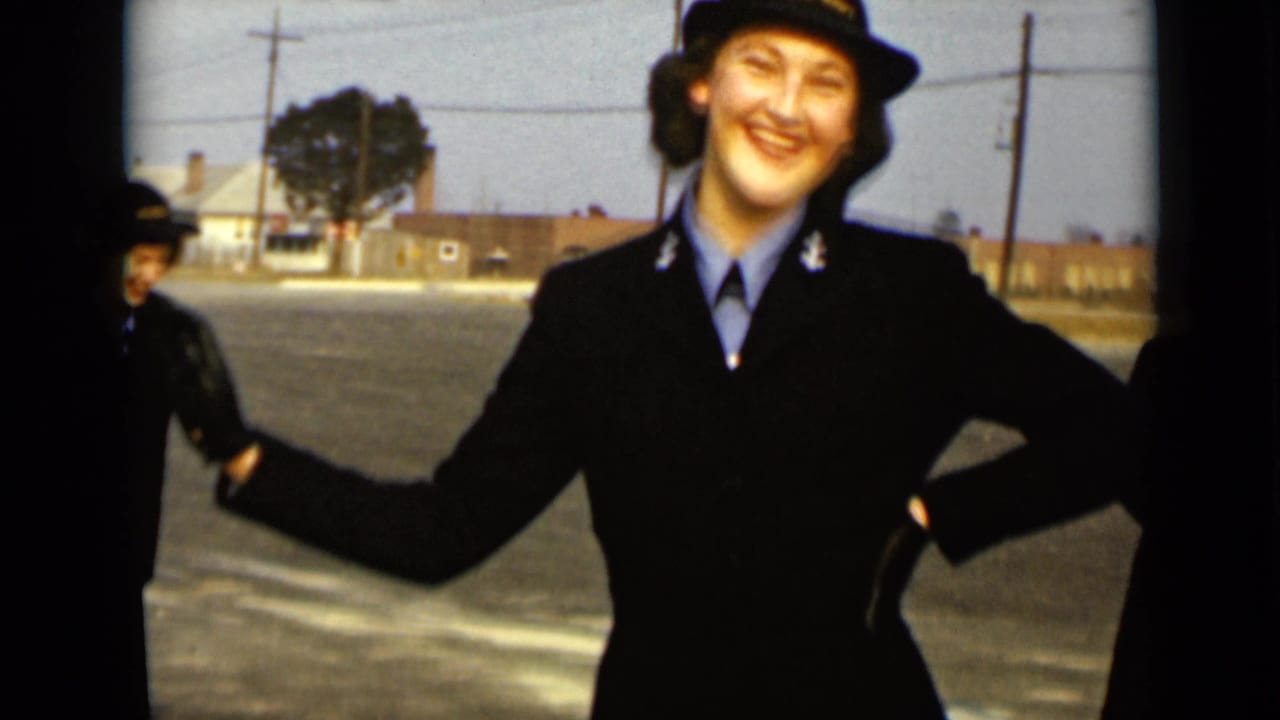 A close view shows a smiling uniformed woman waving in 1947 New York, with another uniformed figure partly visible behind her.