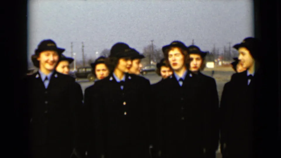 A group of uniformed women stand together on a paved lot in 1947 New York, with mixed smiles and side glances.