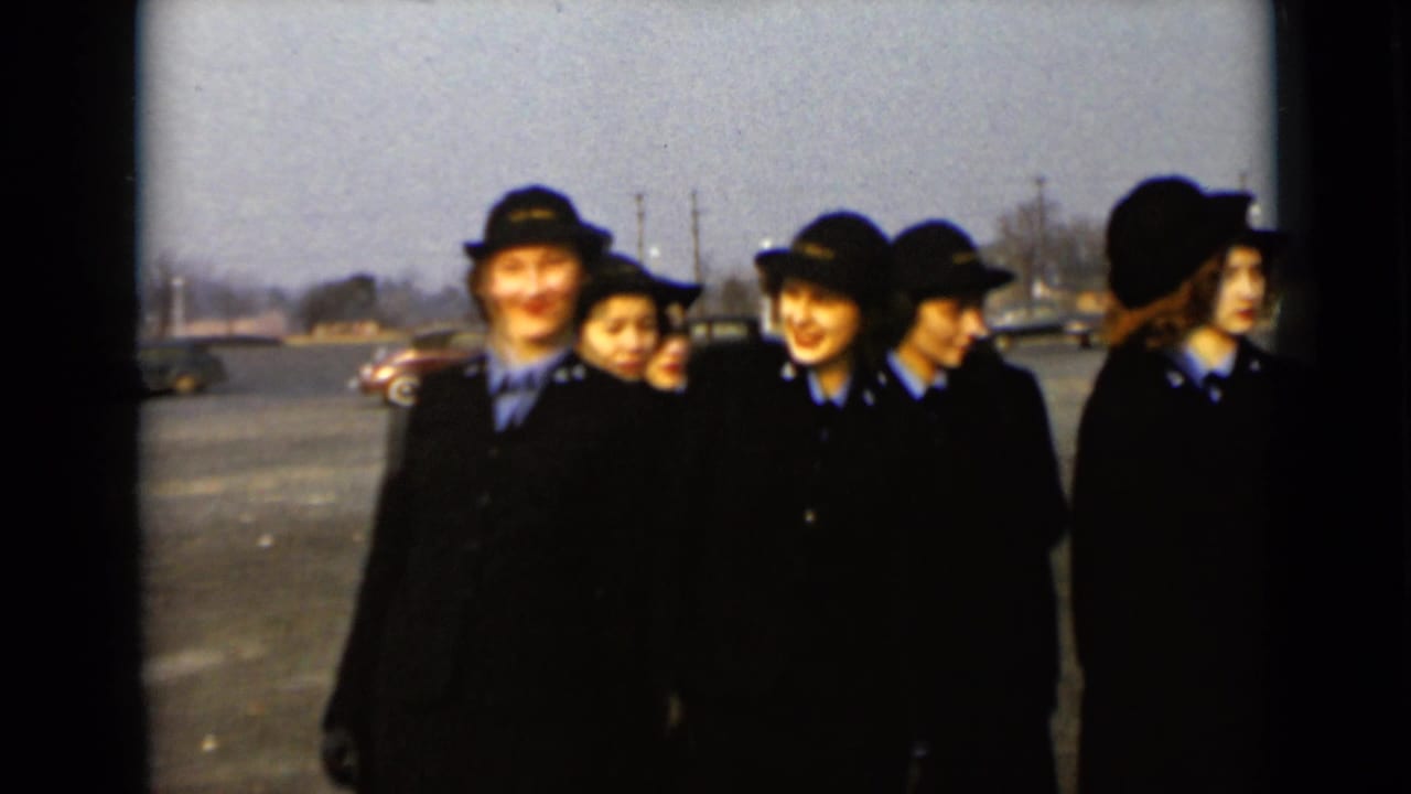 Uniformed women in dark coats and hats stand together in a parking lot.