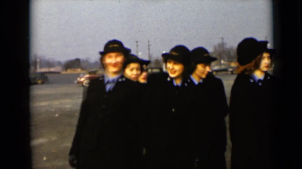 In 1947 New York footage, several uniformed women stand close together outdoors with parked cars and houses behind them.