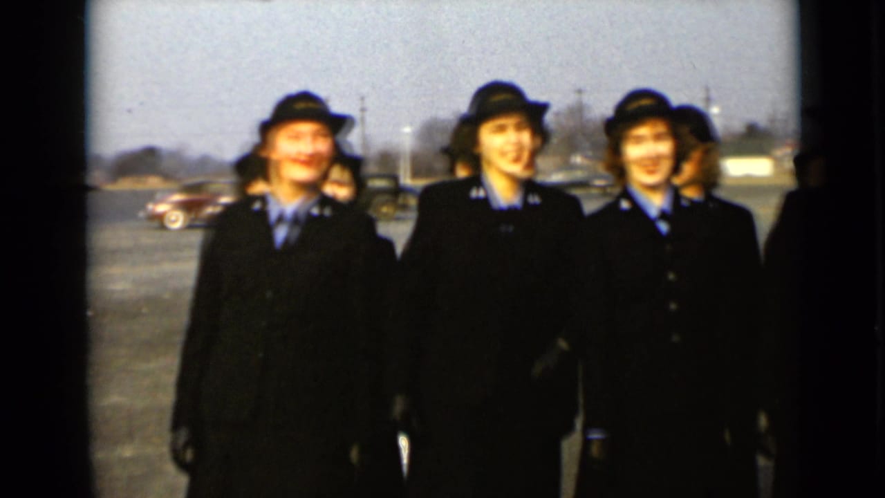 Three uniformed women in dark coats and hats stand outdoors near a paved lot.
