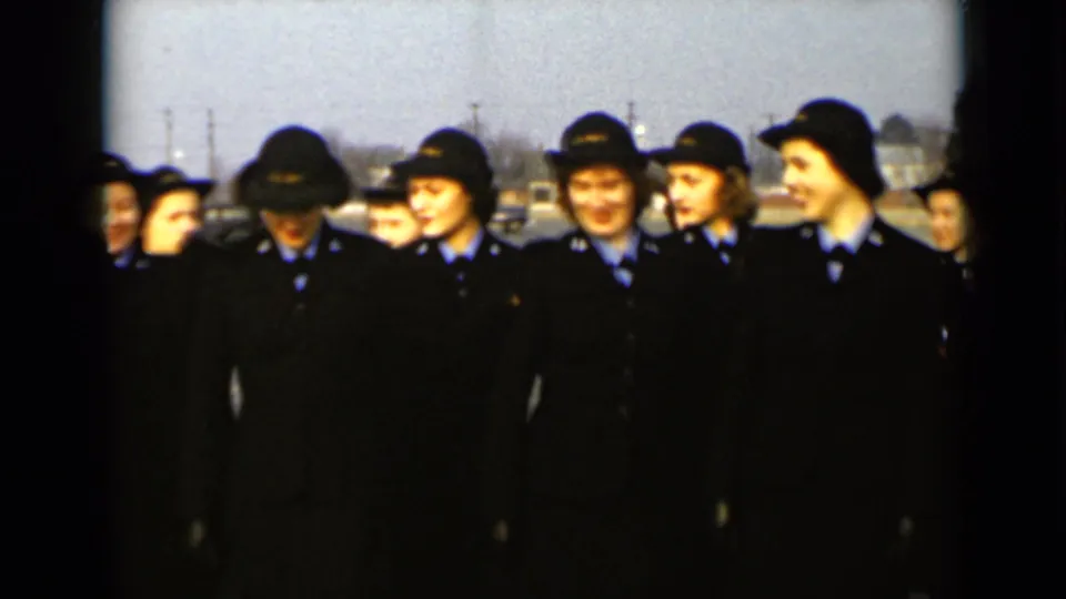 Women in dark formal uniforms and hats stand closely together outdoors in 1947 New York, with a paved lot and distant structures behind them.