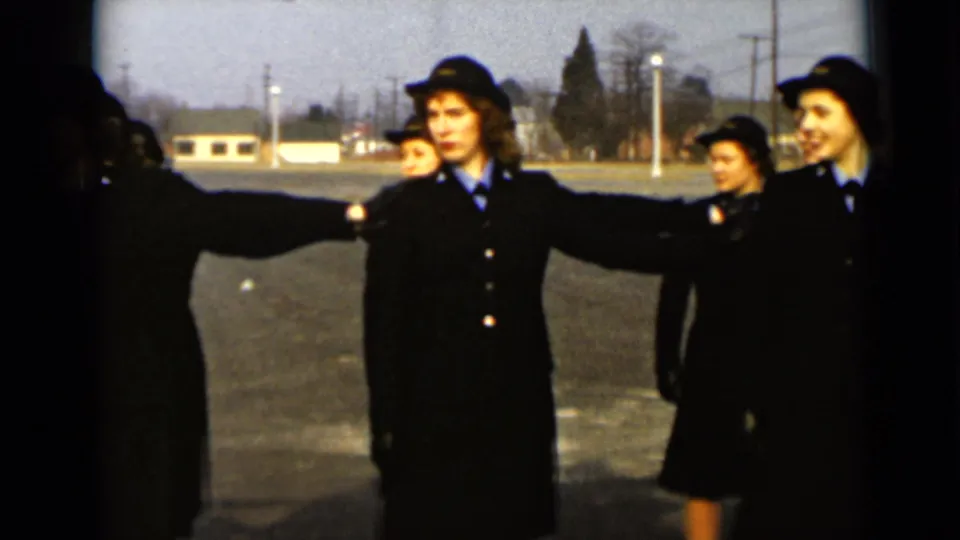 A woman in a dark formal uniform extends her arm as other uniformed women stand nearby in a paved New York outdoor setting from 1947.