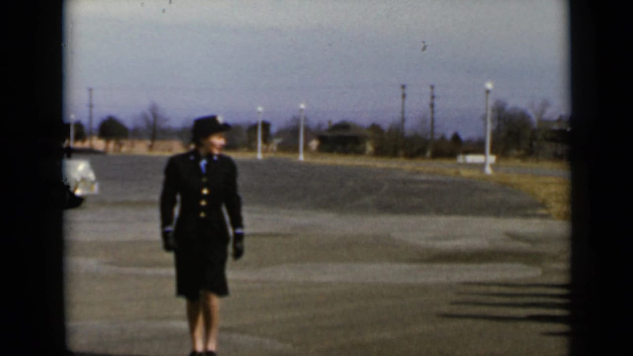 Uniformed woman in a dark suit and hat standing on a paved lot.