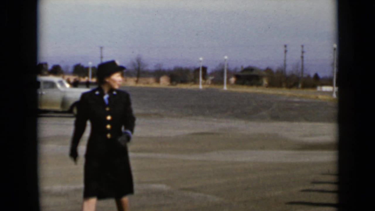Uniformed woman walking across a paved lot near a vintage car.