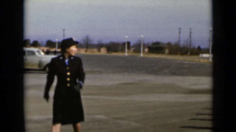 A woman in a dark uniform-style suit and hat walks across a paved lot, with a vintage car behind her.