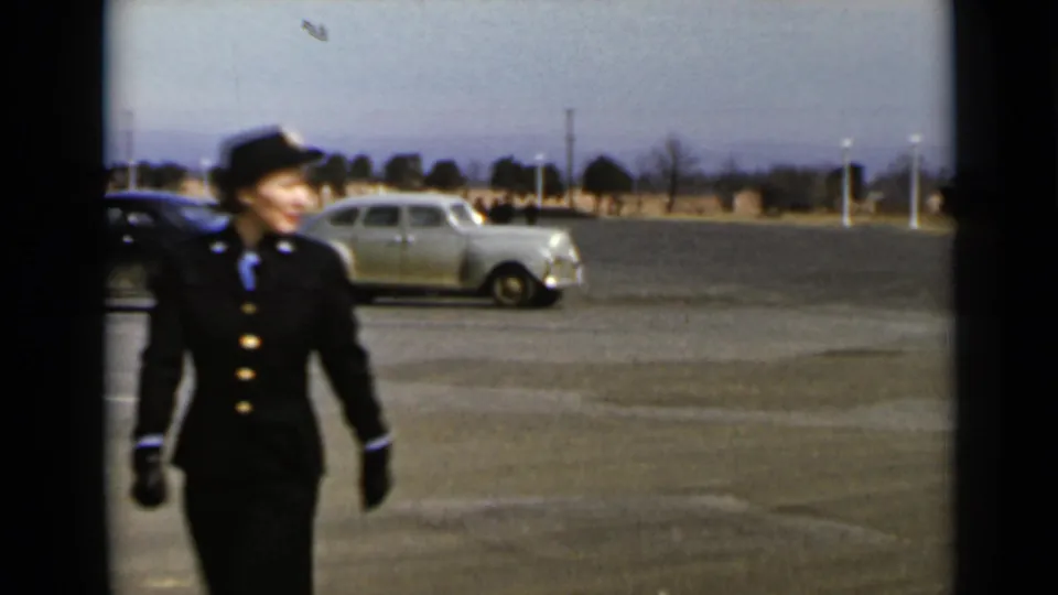 A closer 1947 New York frame shows a woman in a dark uniform-style suit walking past a parked vintage car.