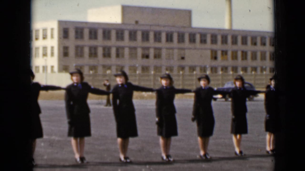 Uniformed women in New York, 1947, hold a synchronized spacing posture in front of a large building and fence.