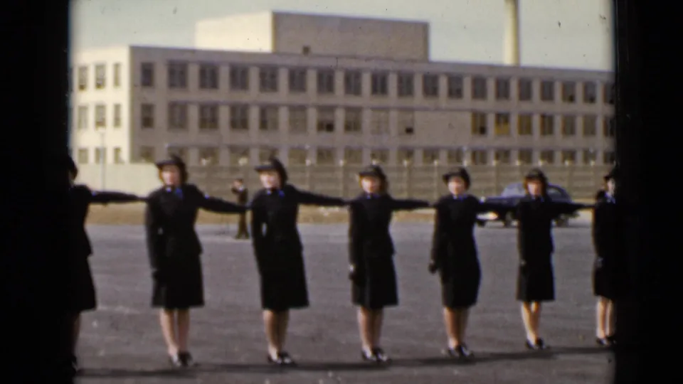 Uniformed women in New York, 1947, hold a synchronized spacing posture in front of a large building and fence.