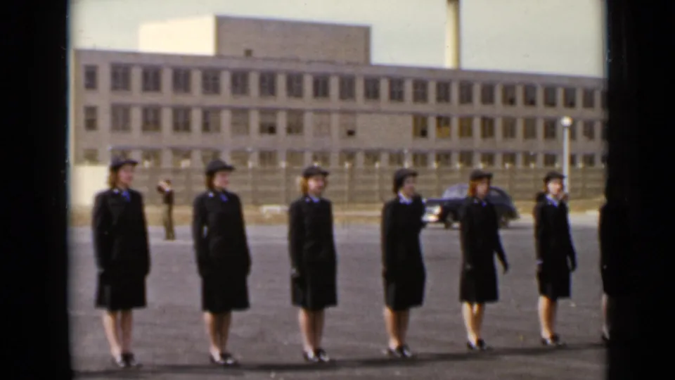 The women transition from arm spacing to attention, with a parked car and institutional building visible behind them.