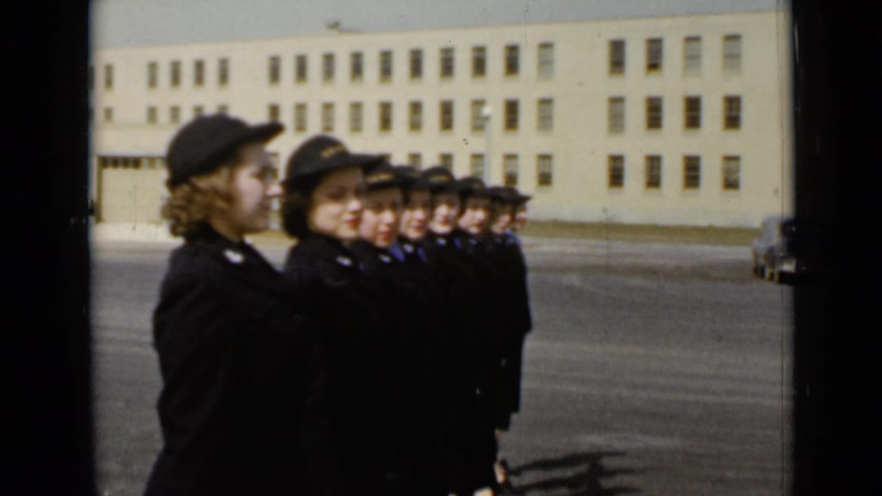 Women in dark uniforms and caps stand in a line outdoors before a large building.