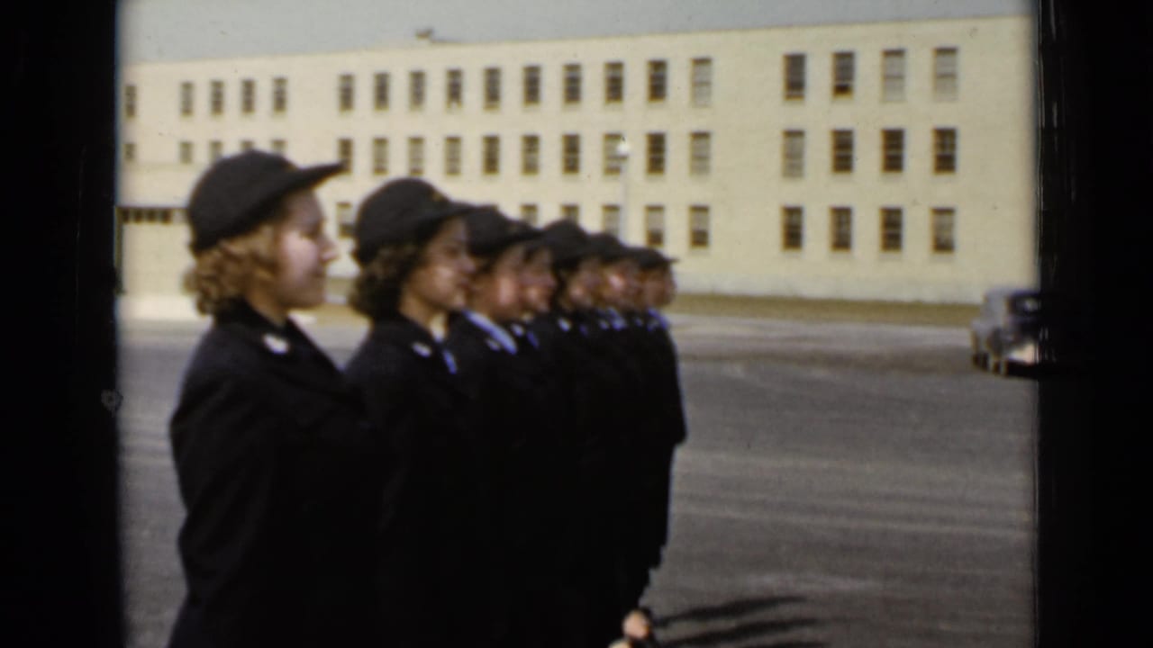 Uniformed women stand in profile, lined up diagonally across a paved outdoor area.