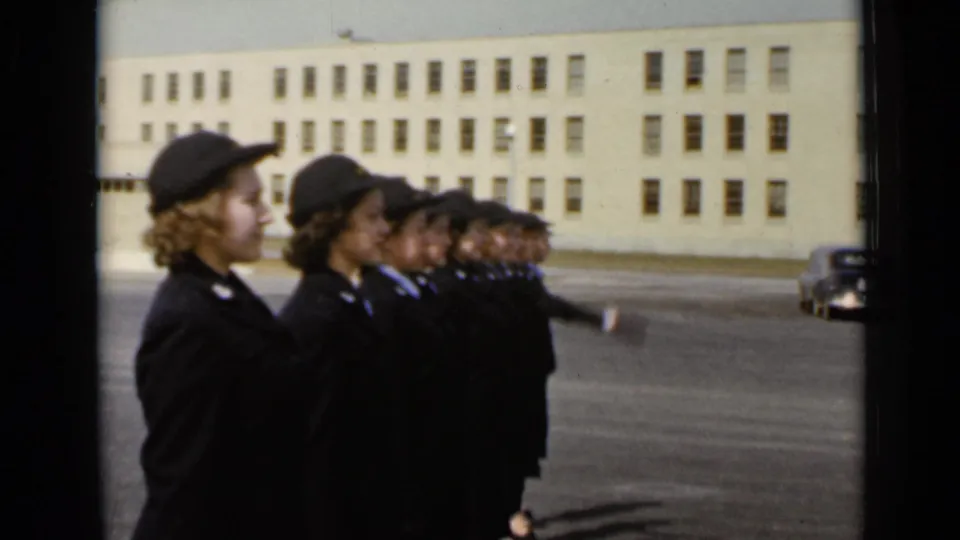 Several uniformed women in the line begin to raise an arm while standing before the large building.