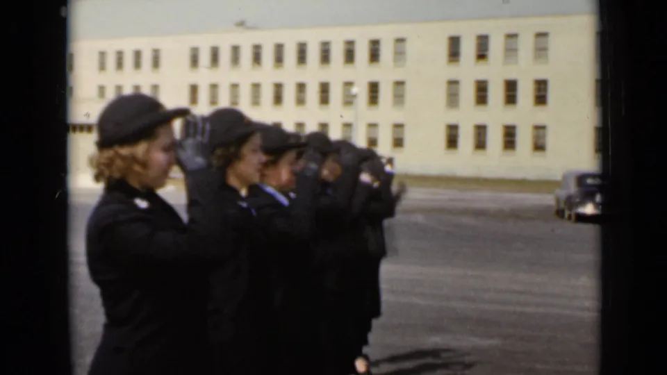 The formation raises hands in a salute-like motion, with faces in profile and a parked car near the building.