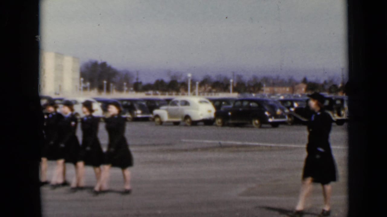 Women in dark uniforms march across a parking lot with vintage cars behind them.