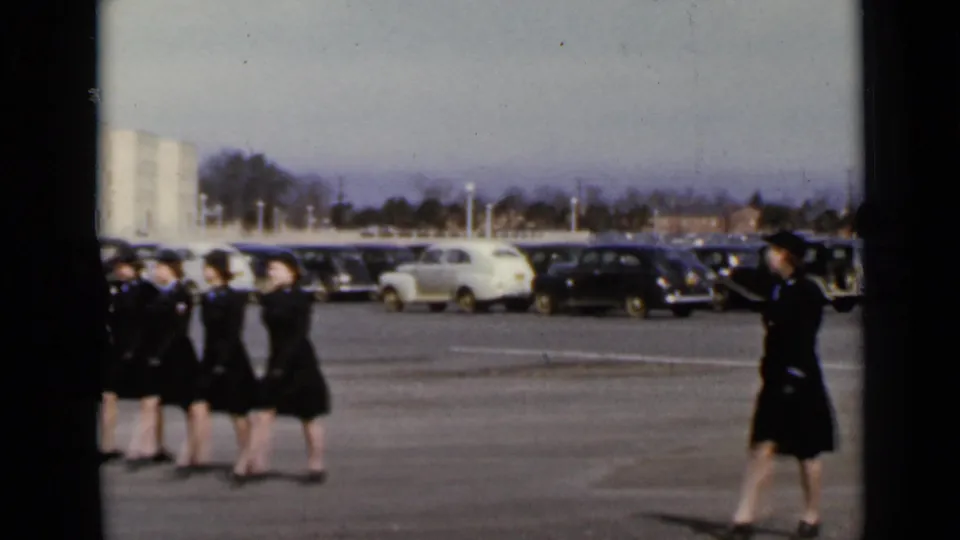Uniformed women march in formation across a New York parking lot in supplied 1947 8mm footage, with parked cars and a light building behind them.