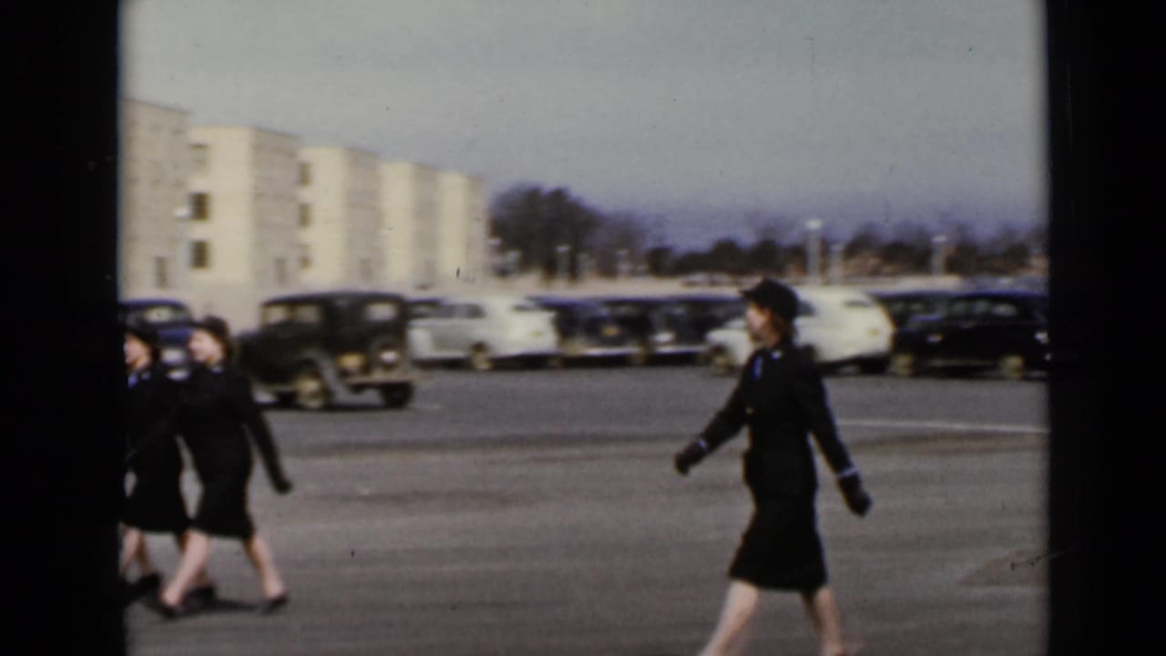 A line of women in dark uniforms marches past parked cars and a light building.