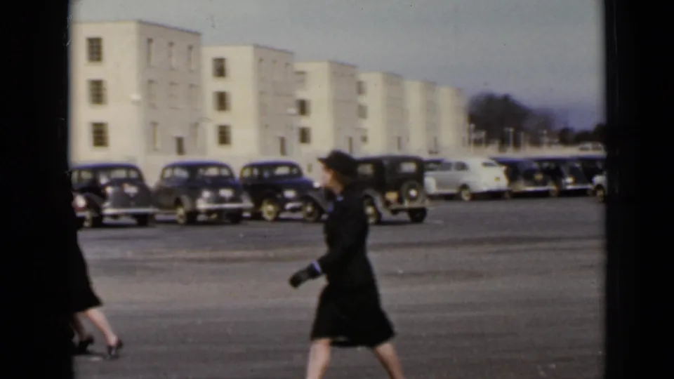 A woman in a dark uniform and hat crosses the frame in the 1947 New York parking-lot footage, with vintage cars and buildings visible.