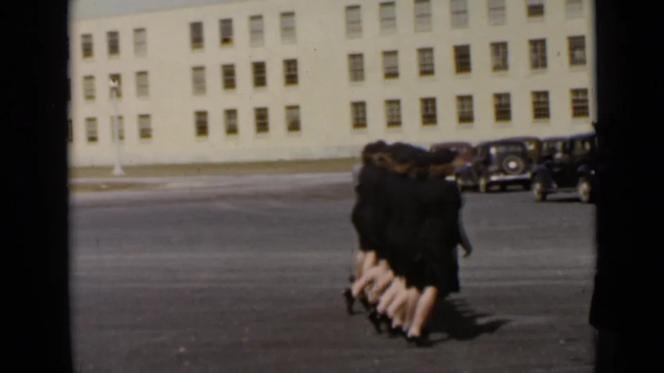 The marching group moves away from the camera in the supplied 1947 New York scene, with a large light-colored building and parked cars beyond.