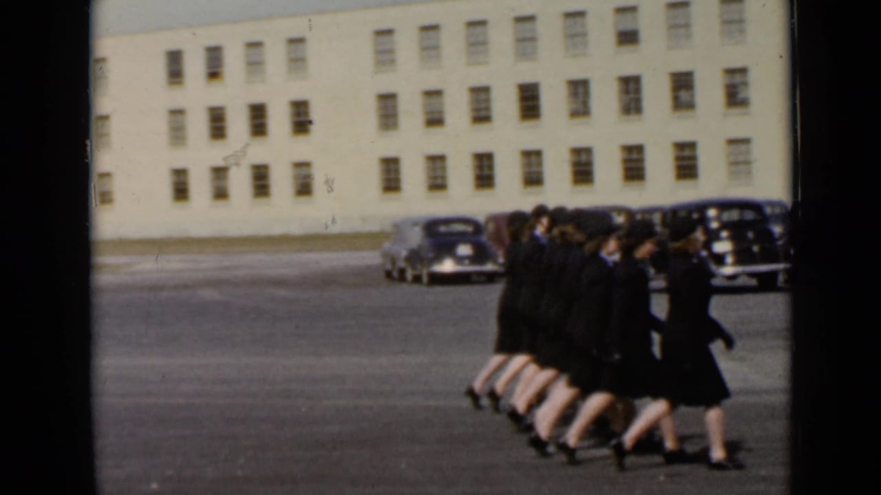 Uniformed women march across a parking lot in front of a pale multi-story building.