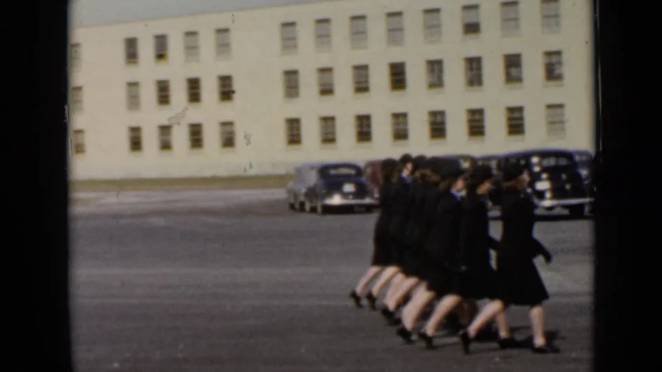 1947 New York 8mm frame showing women in dark uniforms and hats marching in a line near parked mid-century cars.