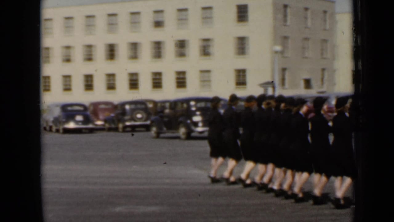 1947 New York frame with a formation of women in matching dark clothing moving past rows of vintage cars.