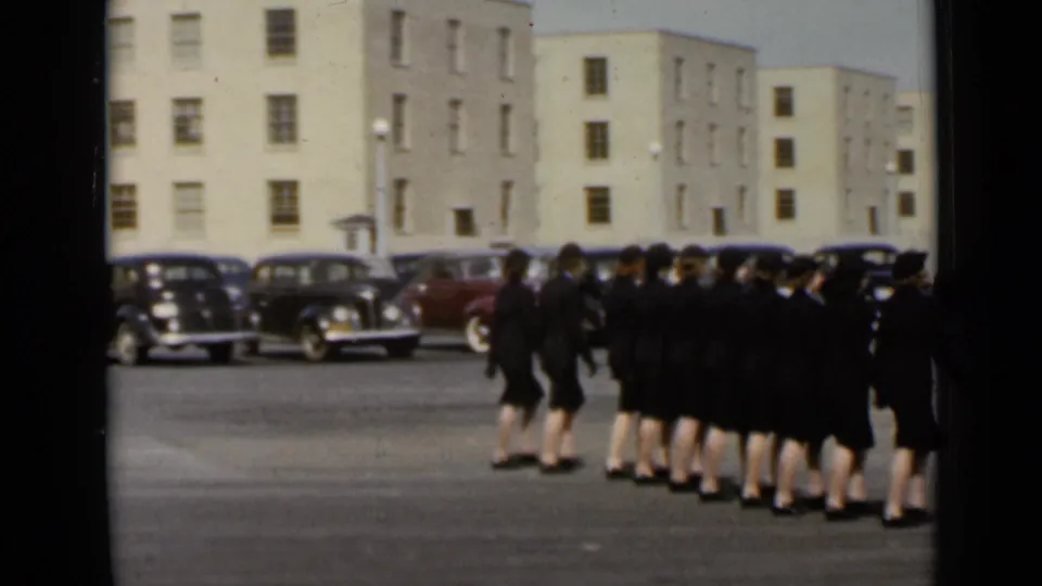 1947 New York 8mm frame showing a group of uniformed women marching in front of mid-century cars and plain buildings.