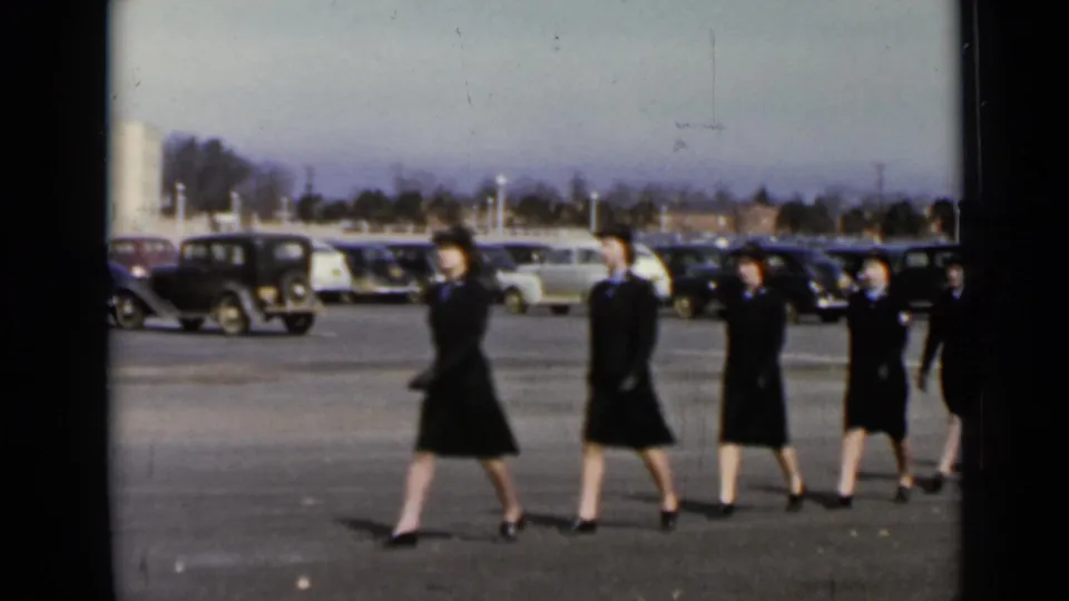 1947 New York frame showing several uniformed women marching in profile across a parking area with cars and distant buildings.