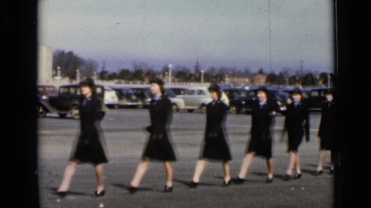 In 1947 New York home-movie footage, women in matching dark uniforms and hats march past parked cars in an open lot.