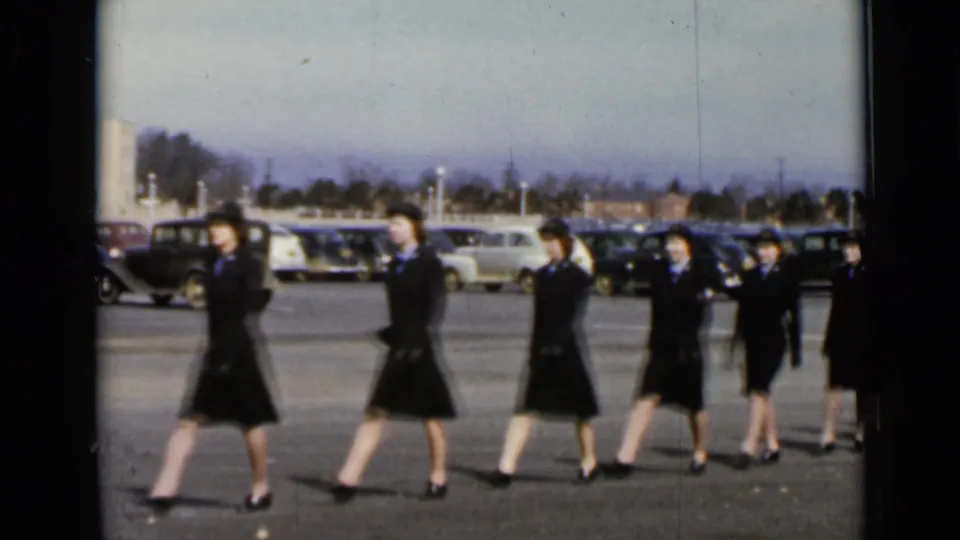 In 1947 New York home-movie footage, women in matching dark uniforms and hats march past parked cars in an open lot.