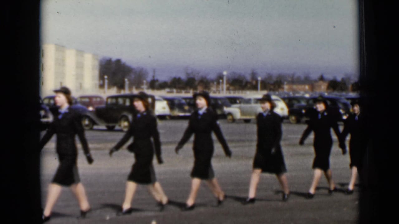Women in matching dark coats and hats continue marching in formation across a New York parking area in 1947 footage.