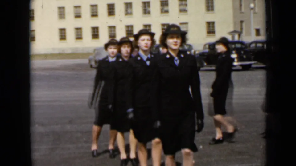 In 1947 New York footage, a line of young women in dark uniforms and hats marches past an institutional building.