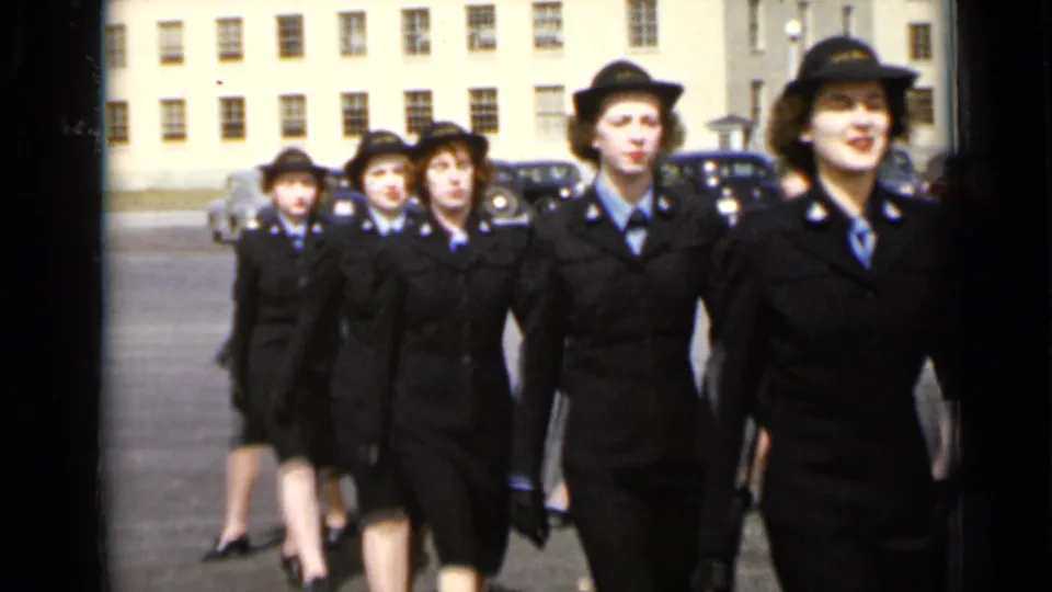 Uniformed women in dark jackets, skirts, ties, and hats march in formation in a New York institutional courtyard.