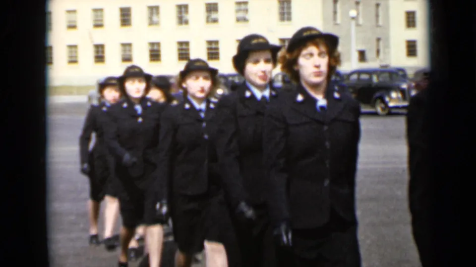 A close view shows women in matching dark uniforms and hats moving in step across the courtyard in 1947 New York.