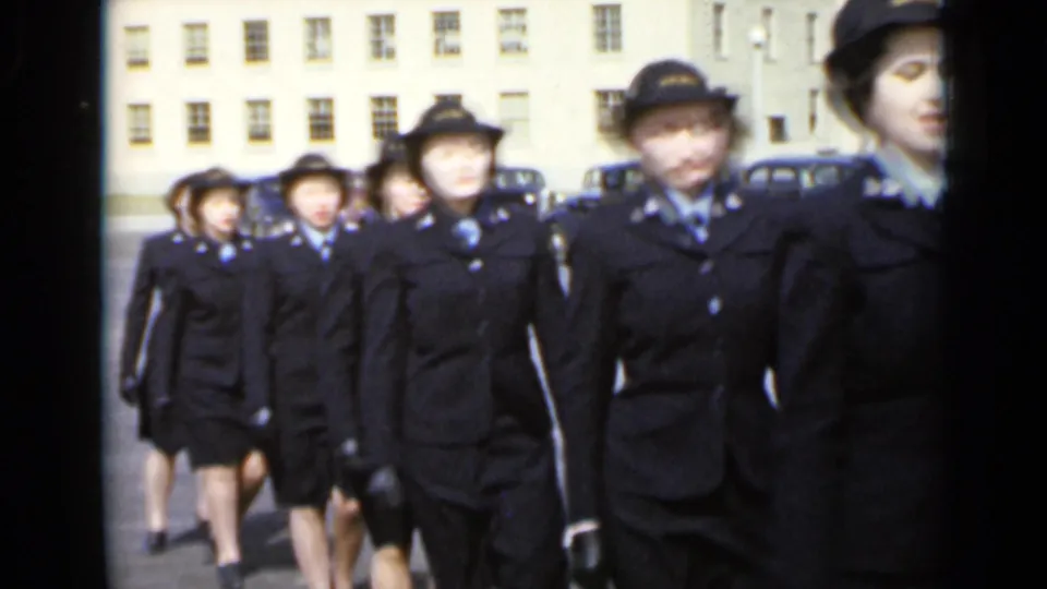Women in matching dark uniforms and hats continue past the handheld camera, with parked cars and a windowed building behind them.